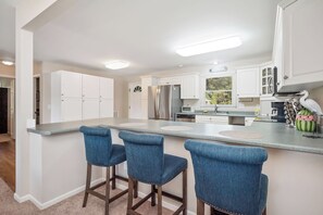 A modern kitchen with white cabinets, a stainless steel refrigerator, and a bar area with blue barstools. - Asleep At The Reel: Waterway Home with Dock (Oak Island)