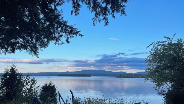 Vista lacustre con una silla y un kayak, rodeado de árboles y cielo despejado.