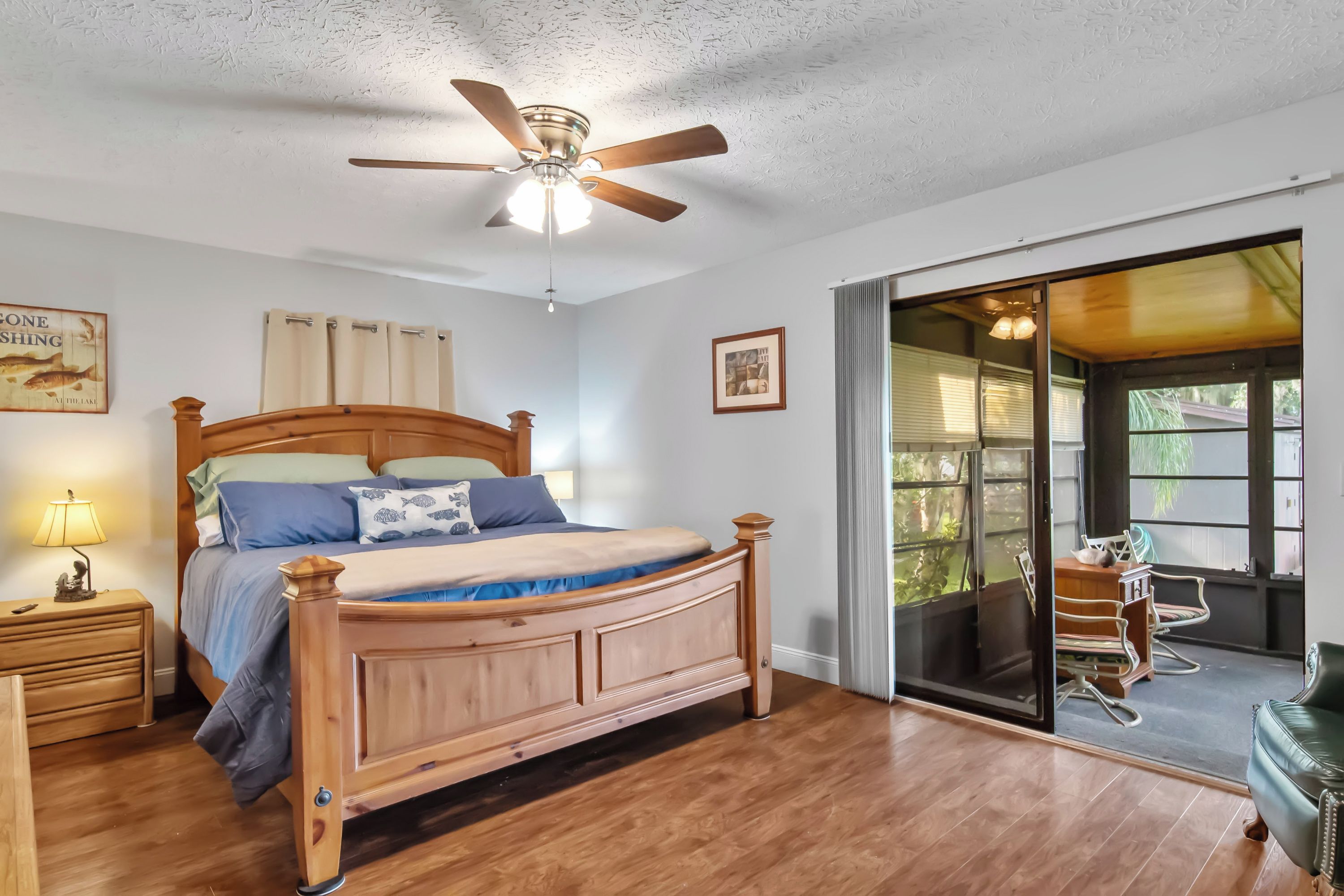 A bedroom with a wooden bed, a ceiling fan, a nightstand with a lamp, and a view of the outdoors through a sliding glass door.