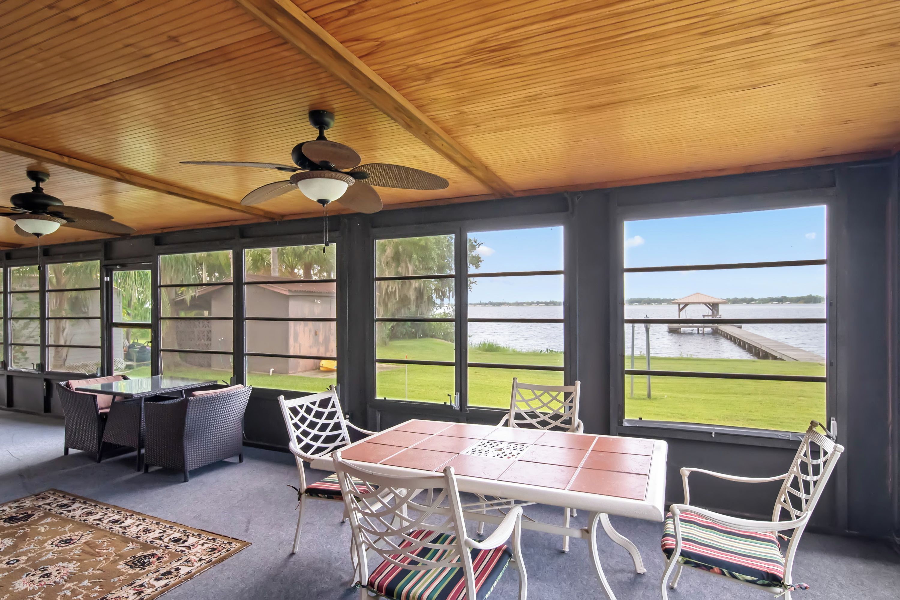 Un área de estar techada al aire libre con una mesa y sillas, ventanas amplias con vistas a un cuerpo de agua y un muelle, y ventiladores de techo.