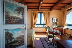 Une pièce avec un grand tableau au mur, une fenêtre avec des rideaux, une table ornée d’un vase de fleurs et une chaise.