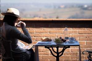 Une personne assise à une table, devant un panier de fruits, une bouteille et une tasse, avec un mur de briques et un paysage en arrière-plan.