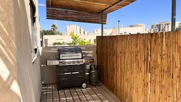 Zona de barbacoa al aire libre con parrilla y basurero, delimitada por una valla de madera y un pérgola.