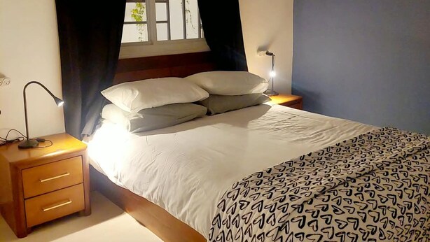 Un dormitorio con cama, mesita de noche, lámpara, ventana con cortinas y pared azul.