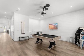A pool table with a ceiling fan, a painting, and a retro arcade machine in a room with white walls and wooden flooring. - Ocean Breeze: Heated Pool & Hot Tub, Free Golf Cart, Two Arcade Rooms (Miramar Beach)