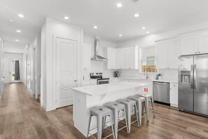 A modern kitchen with white cabinetry, a central island with stools, stainless steel appliances, and a wooden floor. - Ocean Breeze: Heated Pool & Hot Tub, Free Golf Cart, Two Arcade Rooms (Miramar Beach)