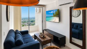A modern living room with a sofa, a coffee table, and a TV. There is a large window with a view of the city.