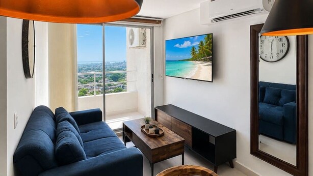 A modern living room with a sofa, a coffee table, and a TV. There is a large window with a view of the city. - Tropical Midnight | Sunset Views & Pool, Wi-Fi (Cartagena)