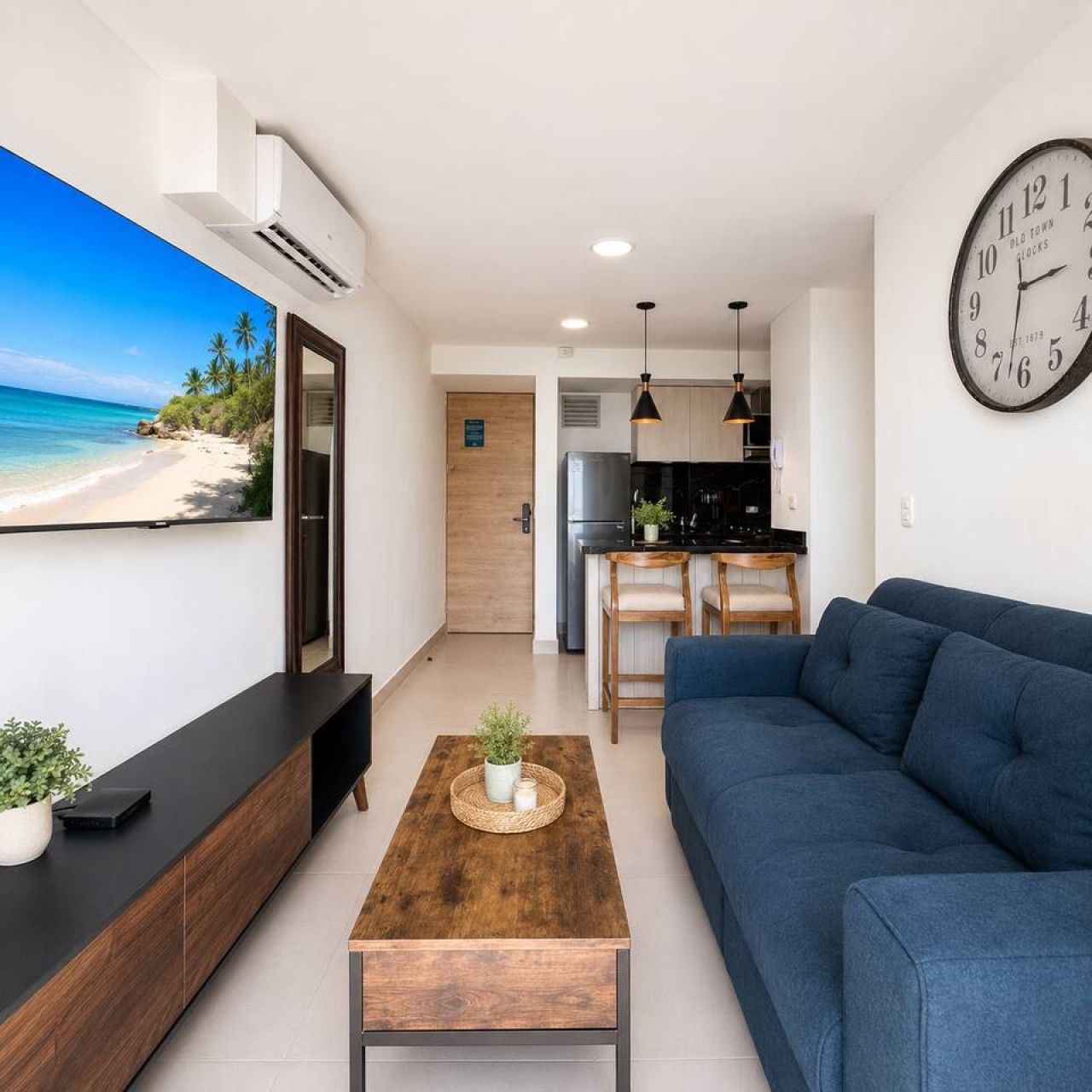A modern living room with a large flat-screen TV, a blue sofa, a wooden coffee table, and a kitchen area in the background.