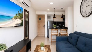 A modern living room with a large flat-screen TV, a blue sofa, a wooden coffee table, and a kitchen area in the background. - Tropical Midnight | Sunset Views & Pool, Wi-Fi (Cartagena)