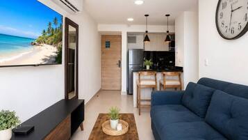 A modern living room with a large flat-screen TV, a blue sofa, a wooden coffee table, and a kitchen area in the background.