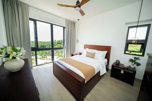 Ein Schlafzimmer mit einem großen Bett, einem Nachttisch, einem Wendetisch, einem Deckenventilator und einem Fenster mit Blick ins Grüne.