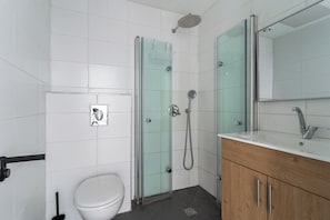 A bathroom with a toilet, a glass shower enclosure, a sink with a mirror, and a wooden cabinet. - Blueground - Historical Preserved Estate (Tel Aviv-Yafo)