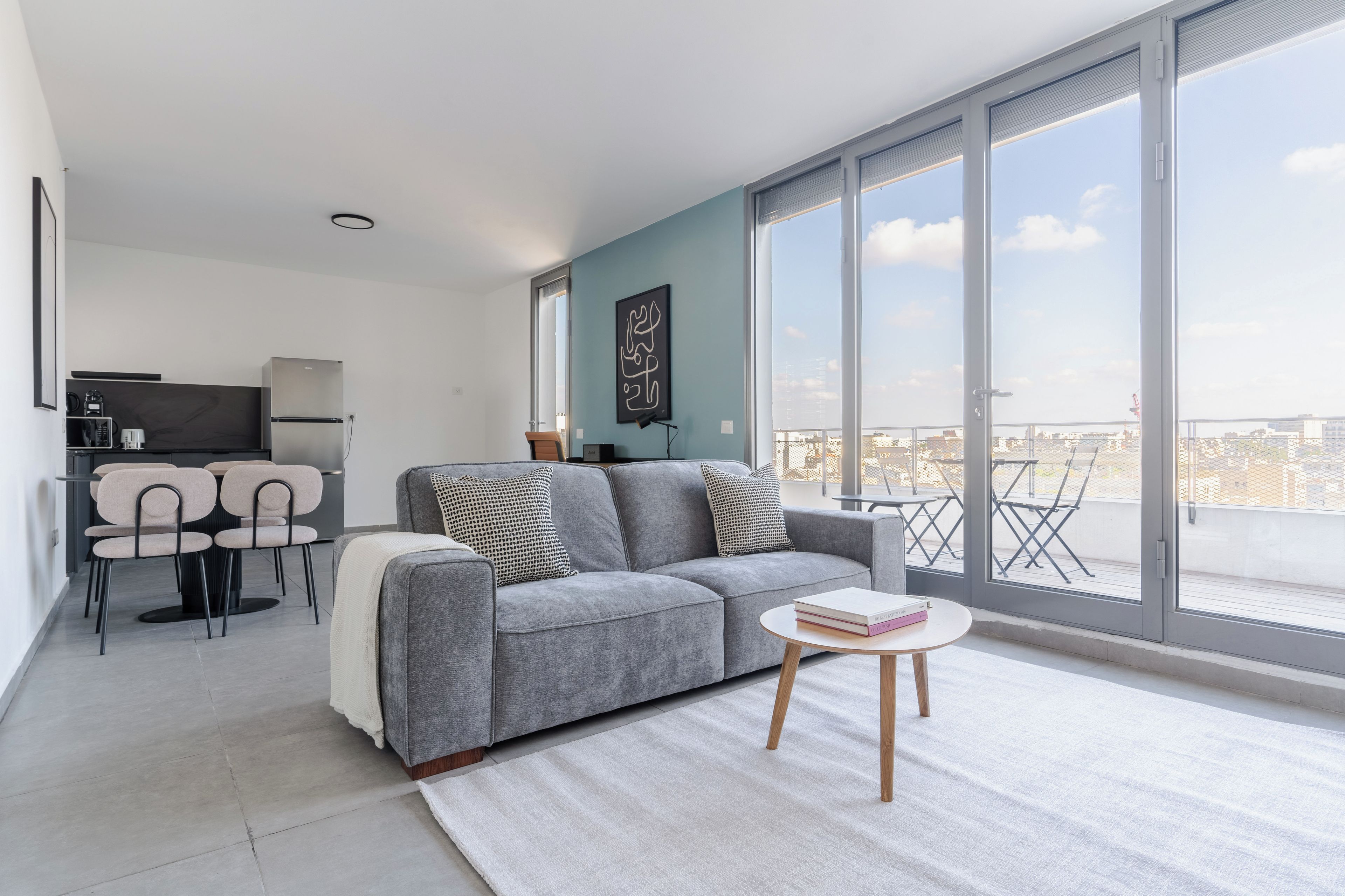 Una sala moderna con un sofá gris, una mesita redonda con libros y vistas a la ciudad a través de grandes ventanales.