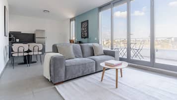 Una sala moderna con un sofá gris, una mesita redonda con libros y vistas a la ciudad a través de grandes ventanales.