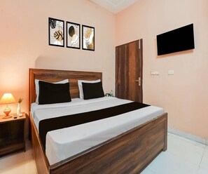 A neatly made bed with a wooden headboard, a bedside table with a lamp, and a wall-mounted TV. - Collection O City Centre Sapror (Phagwara)