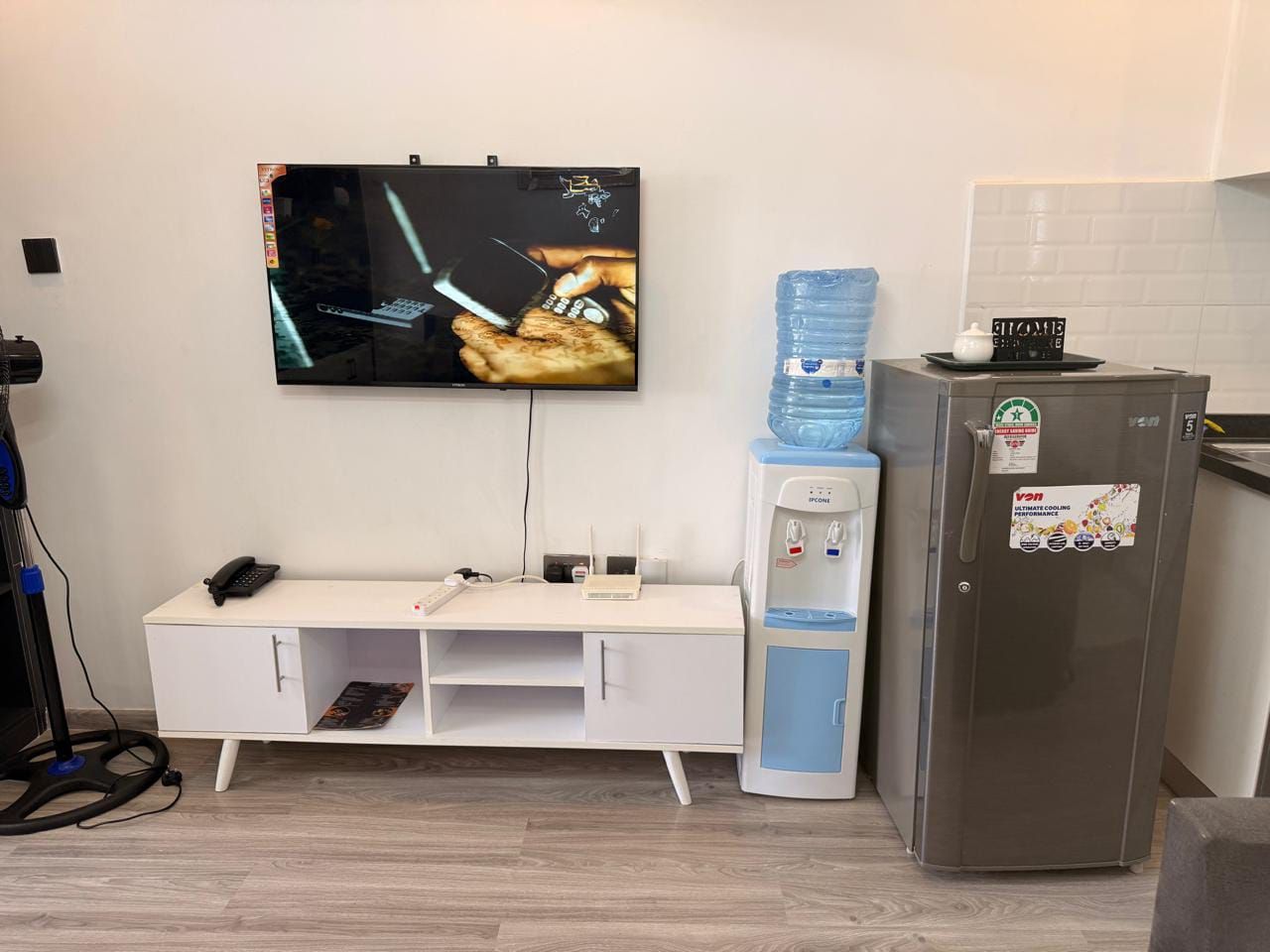 Une petite cuisine équipée d’un réfrigérateur, d’un distributeur d’eau, d’un téléviseur à écran plat, d’un bureau avec un téléphone et d’un système de gestion des câbles fixé au mur.