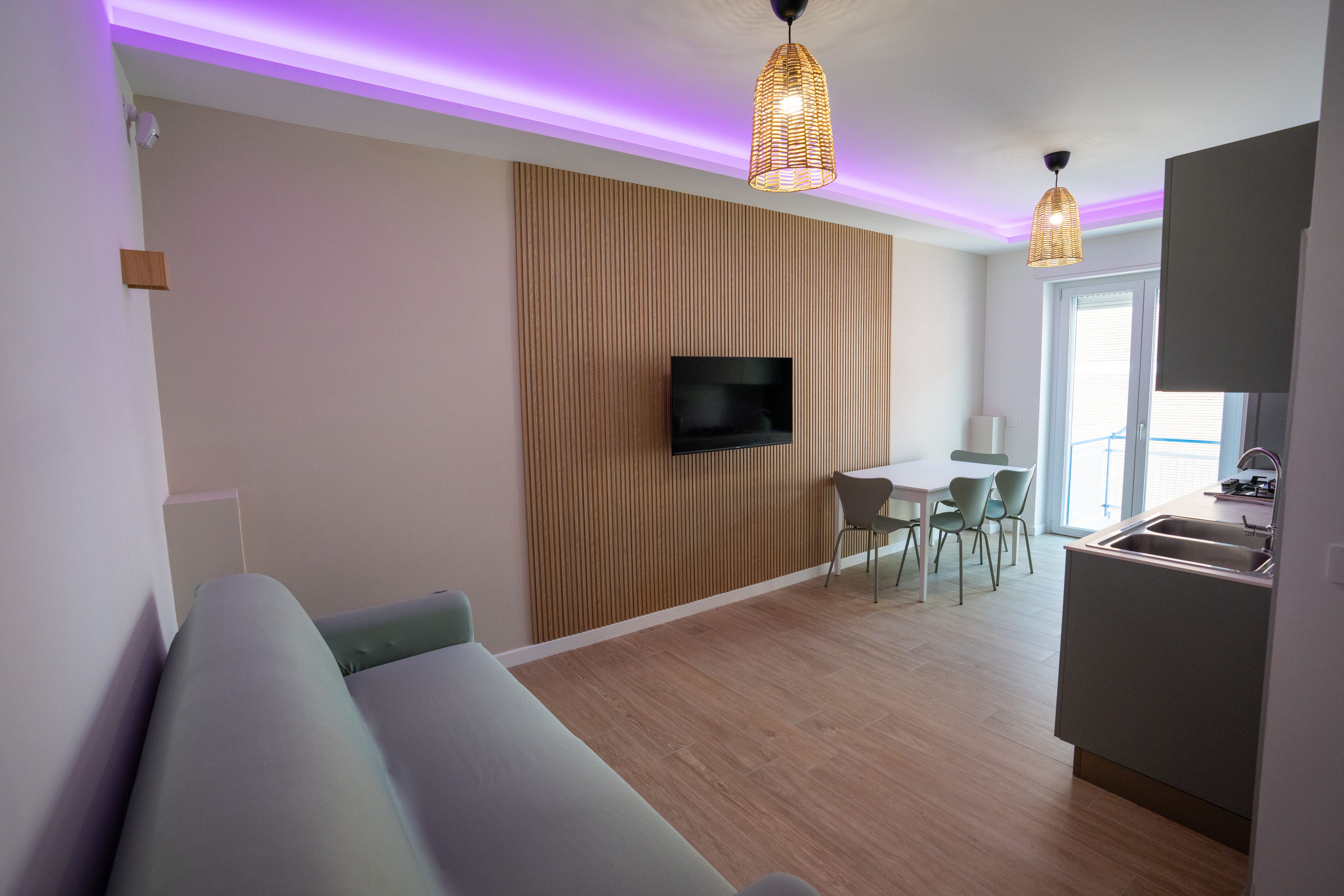 Un salon moderne doté d’un téléviseur à écran plat fixé sur un mur recouvert de panneaux en bois, d’un coin repas avec une table et des chaises, et d’une cuisine équipée d’un évier et d’une cuisinière.