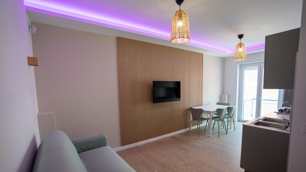 Ein modernes Wohnzimmer mit einem Flachbildfernseher an einer Wand aus Holzpaneelen, einem Essbereich mit Tisch und Stühlen sowie einer Küche mit Spüle und Herd.