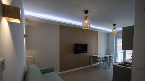 Ein modernes Wohnzimmer mit einer Couch, einem Essbereich mit Stühlen, einem an der Wand montierten Fernseher und einer Küchenzeile mit Spüle.