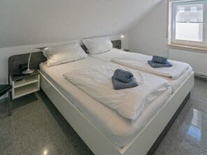 A twin bed with white linens and a grey blanket, a bedside table with a lamp, and a window with blinds. - Holiday Apartment With 1 Bedroom (Esens)