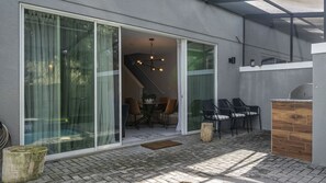 A modern patio area with a dining table and chairs, a wooden bench, and a large sliding glass door leading to an interior space. - Prime Location 4BR Le Reve with Pool Near Disney (Kissimmee)