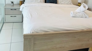 A hotel room with a large bed, two bedside tables, a nightstand, a small potted plant, a painting on the wall, and a towel on the bed. - Phil's Properties At Embassy Gardens GH (Accra)