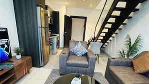 A modern living room with a sofa, armchair, and coffee table. There is a staircase with a wooden railing and a kitchen area with a refrigerator and washing machine. - Phil's Properties At Embassy Gardens GH (Accra)