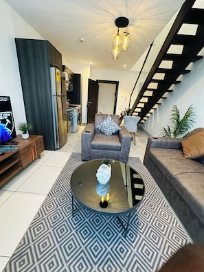 A modern living room with a sofa, armchair, and coffee table. There is a staircase with a wooden railing and a kitchen area with a refrigerator and washing machine. - Phil's Properties At Embassy Gardens GH (Accra)