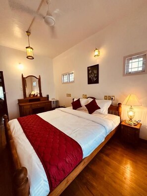 A bedroom with a wooden bed, a nightstand with a lamp, a ceiling fan, and a window with blinds. - Cleety's by MGDC (Kochi)