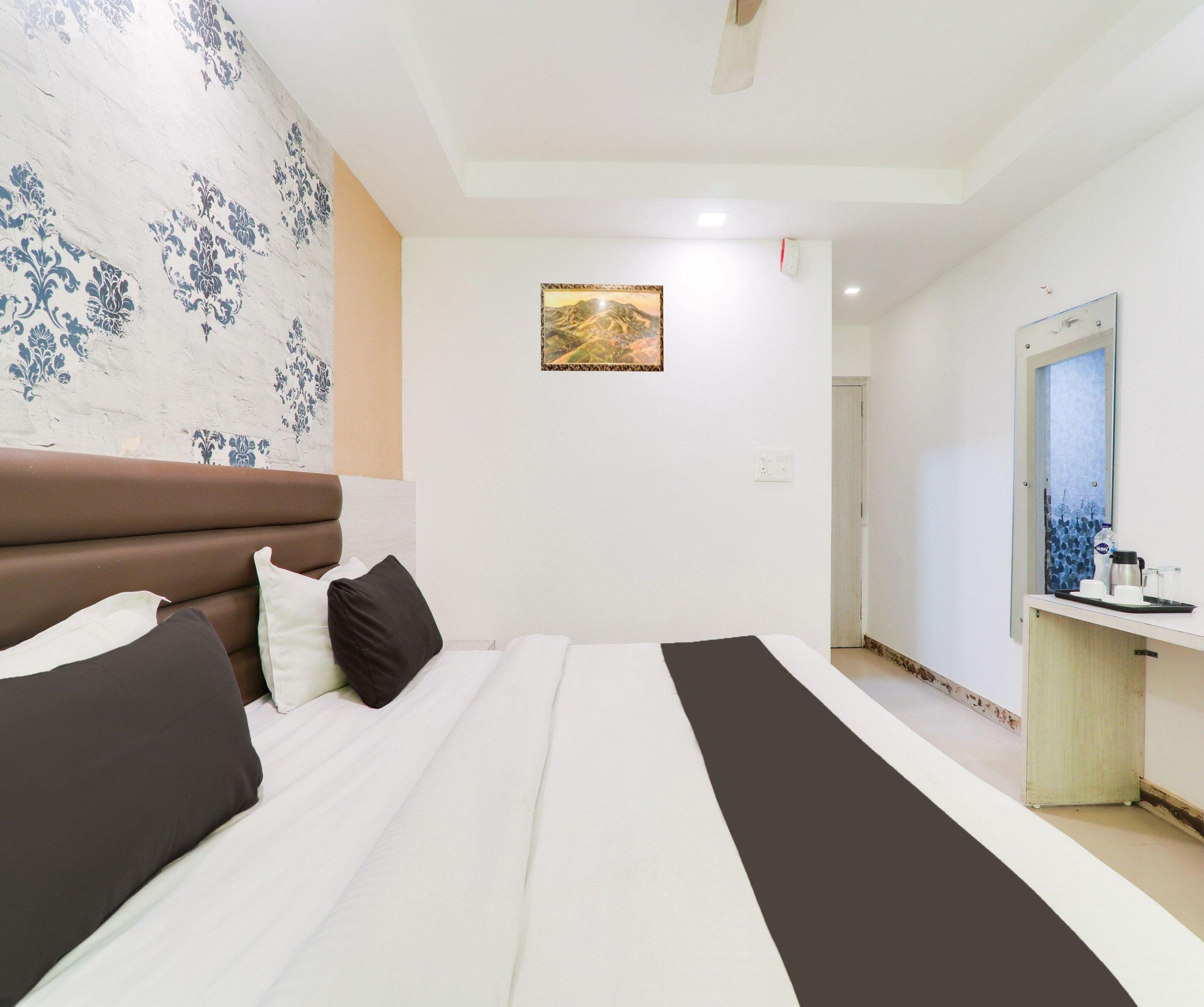A bedroom with a bed, pillows, a headboard, a wall with a floral pattern, a painting, a mirror, and a desk with a tray.