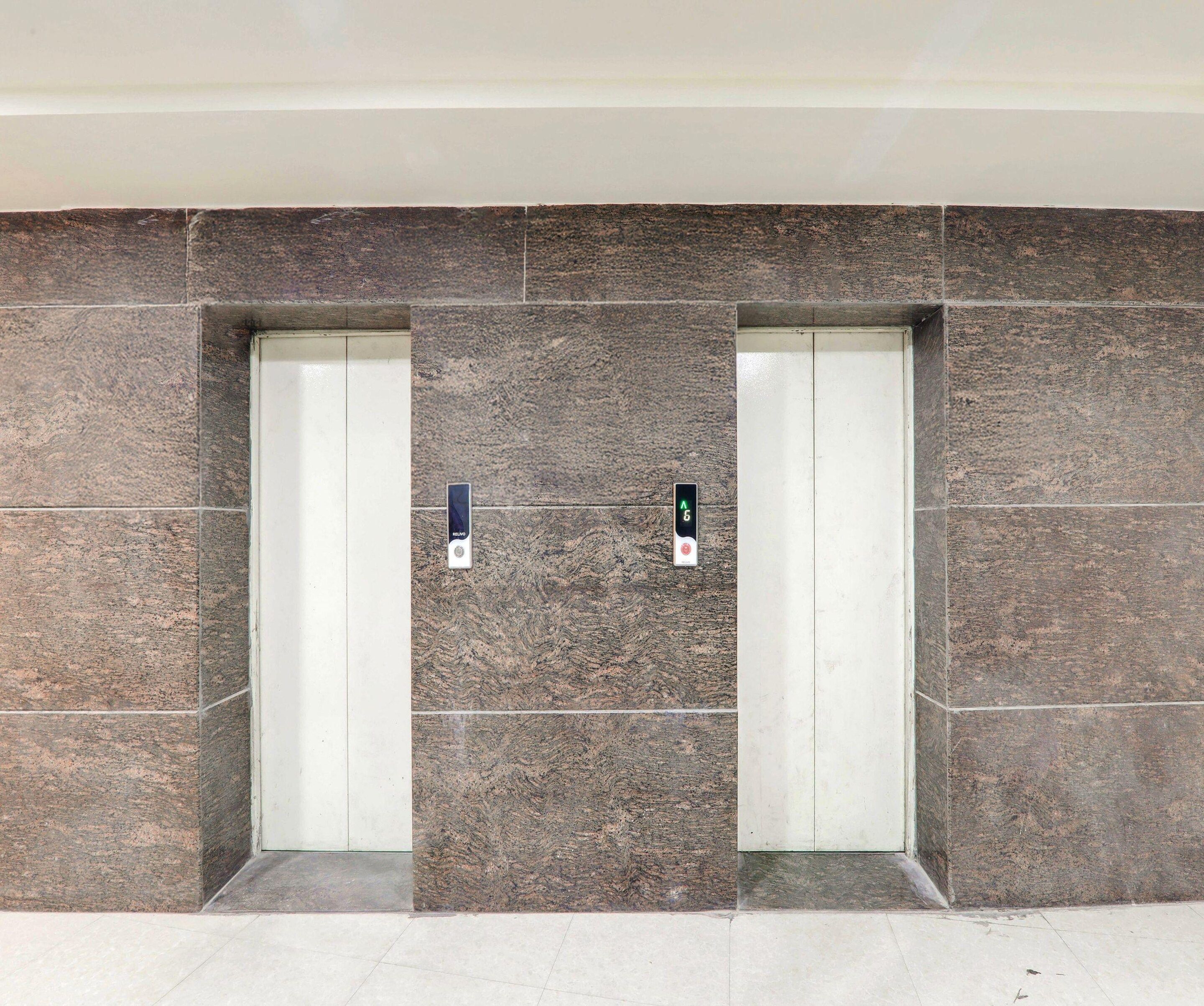 Two elevator doors with a textured wall and a floor with a light-colored tile pattern.