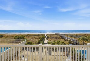 Une vue sur la plage depuis un balcon avec une terrasse en bois, une piscine et une promenade qui mène à la plage.