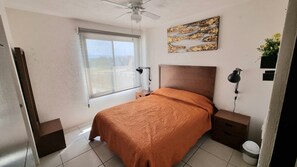 A bedroom with a bed, a window, a ceiling fan, a bedside table, a nightstand, and a wall-mounted lamp. - Casa Carmin (El Guaje)