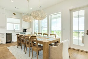 Een moderne keuken met witte kasten, een houten eettafel en uitzicht op de oceaan door grote ramen.