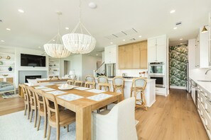 A spacious dining area with a wooden table and chairs, a modern kitchen with white cabinetry, and a large chandelier. - Getaway to 7 Saturdays - Beachside! Beautiful New Construction 2nd Row Home (Galveston)