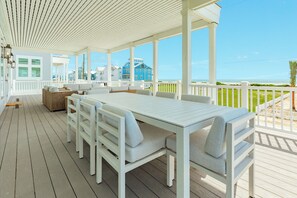 Een overdekt terras met een witte tafel en stoelen, met uitzicht op een strand en huizen.