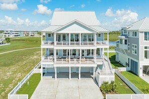 A large, multi-story residential building with a prominent balcony on the second floor, a covered front porch, and a driveway leading to a gated entrance. - Getaway to 7 Saturdays - Beachside! Beautiful New Construction 2nd Row Home (Galveston)