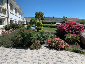 A paved courtyard with a variety of flowering plants and shrubs, a parked car, and a building with a white facade in the background. - LuciRose House: Large 150-square-meter House Near the Cattenom Power Plant – 4 Bedrooms, 2 Kitchens (Cattenom)
