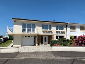 A two-story residential building with a garage, a balcony, and a flower bed with pink flowers. - LuciRose House: Large 150-square-meter House Near the Cattenom Power Plant – 4 Bedrooms, 2 Kitchens (Cattenom)
