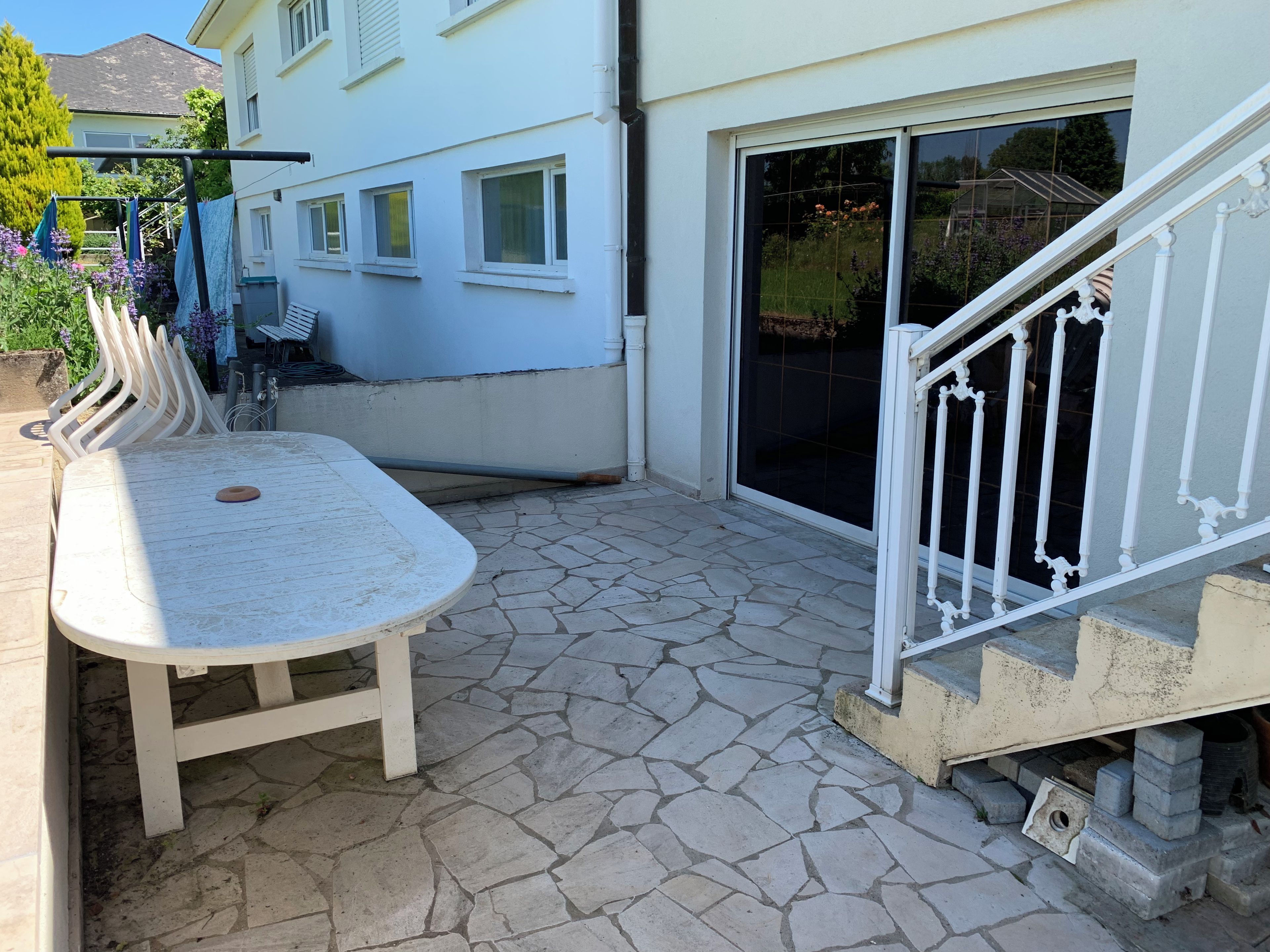 Une terrasse avec une table et des chaises, un sol en pierre, une balustrade blanche et un bâtiment doté de fenêtres et d’une porte coulissante.