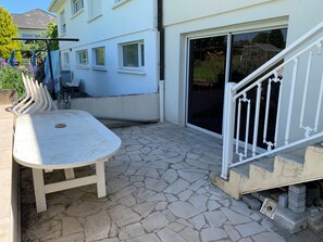 A patio with a table and chairs, a stone floor, a white railing, and a building with windows and a sliding door.