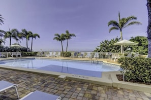 A swimming pool with lounge chairs and umbrellas, surrounded by palm trees and greenery. - Ground Floor, 3-bedroom condo in Indian Rocks Beach, Sunset Paradise, Gulf Side (Indian Rocks Beach)
