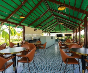 An indoor dining area with round tables and orange chairs, a tiled floor, and a green roof structure.