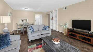 Ein Wohnzimmer mit grauem Sofa, einem Couchtisch mit einer Schale Snacks, einem Flachbildfernseher und einem Essbereich mit einem weißen Schrank und einem Spiegel.