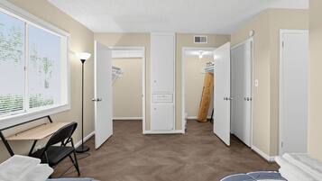 Quarto com escrivaninha, cadeira e uma grande janela. Há portas que dão para outros cômodos e um armário com roupas penduradas.