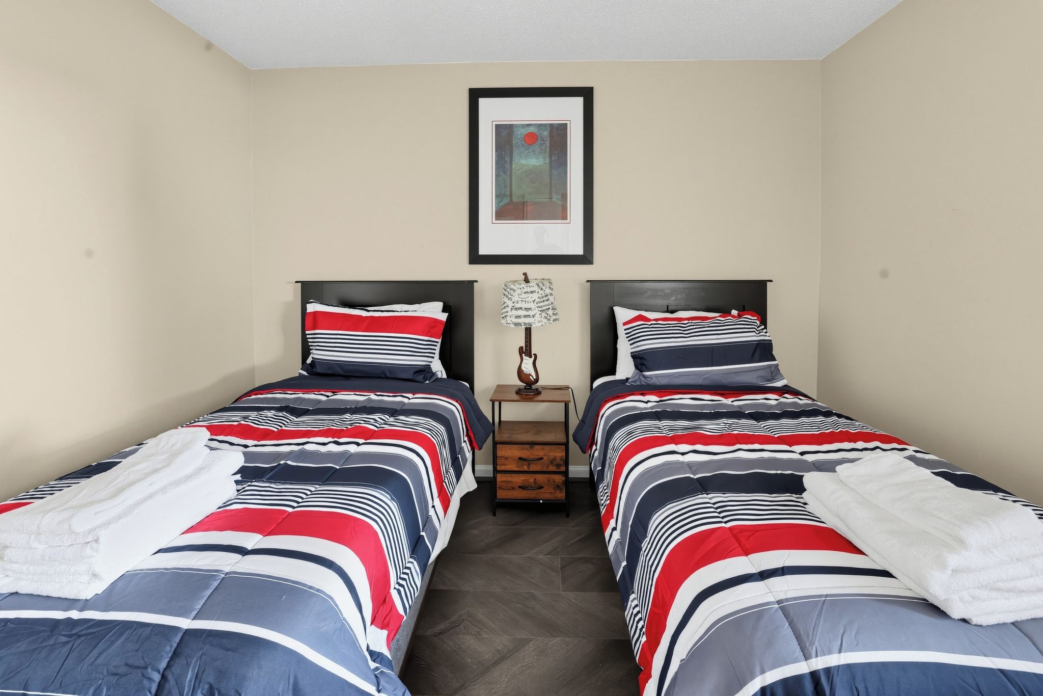 Une chambre double avec deux lits, chacun doté de draps à rayures, une petite table avec une lampe et un tableau encadré au mur.