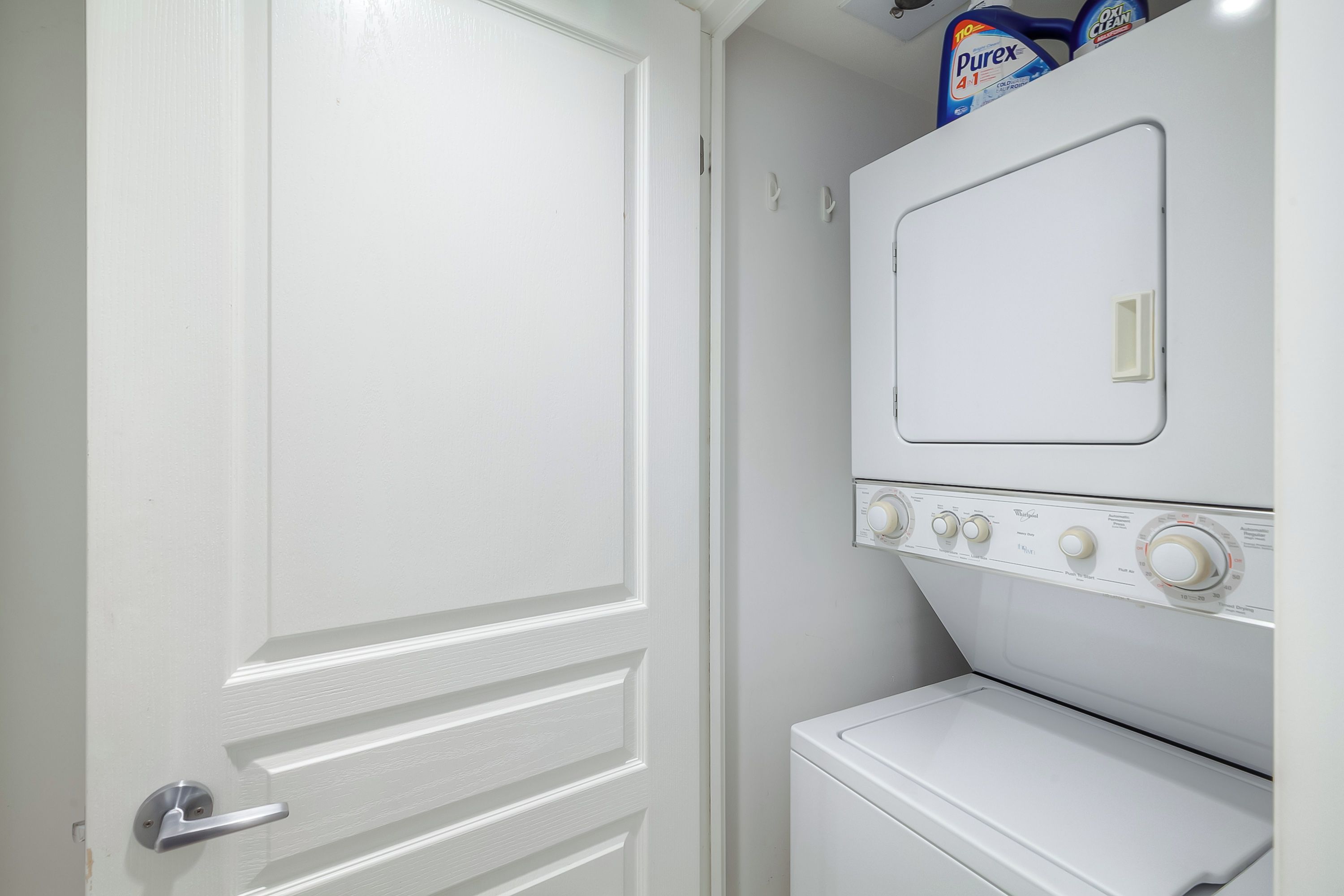 Un lavadero con una lavadora y secadora blancas, una puerta con picaporte plateado y un estante con detergente para la ropa.