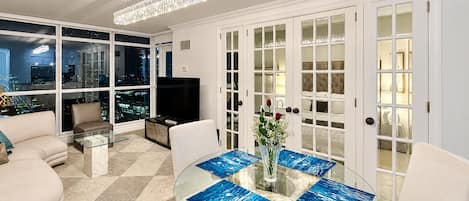 Una sala de estar moderna con una mesa de centro de vidrio, sofás, un televisor y vistas a la ciudad a través de amplias ventanas.