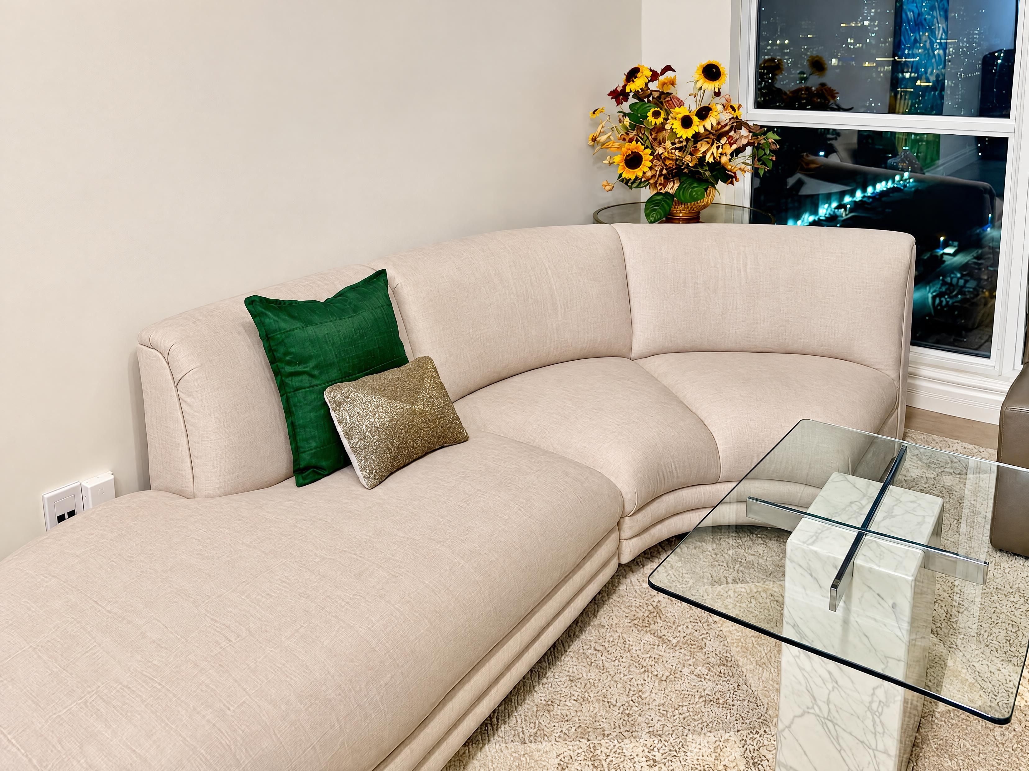 Un sofá color beige con un cojín verde y dorado, una mesa de centro de cristal y un jarrón con flores secas.